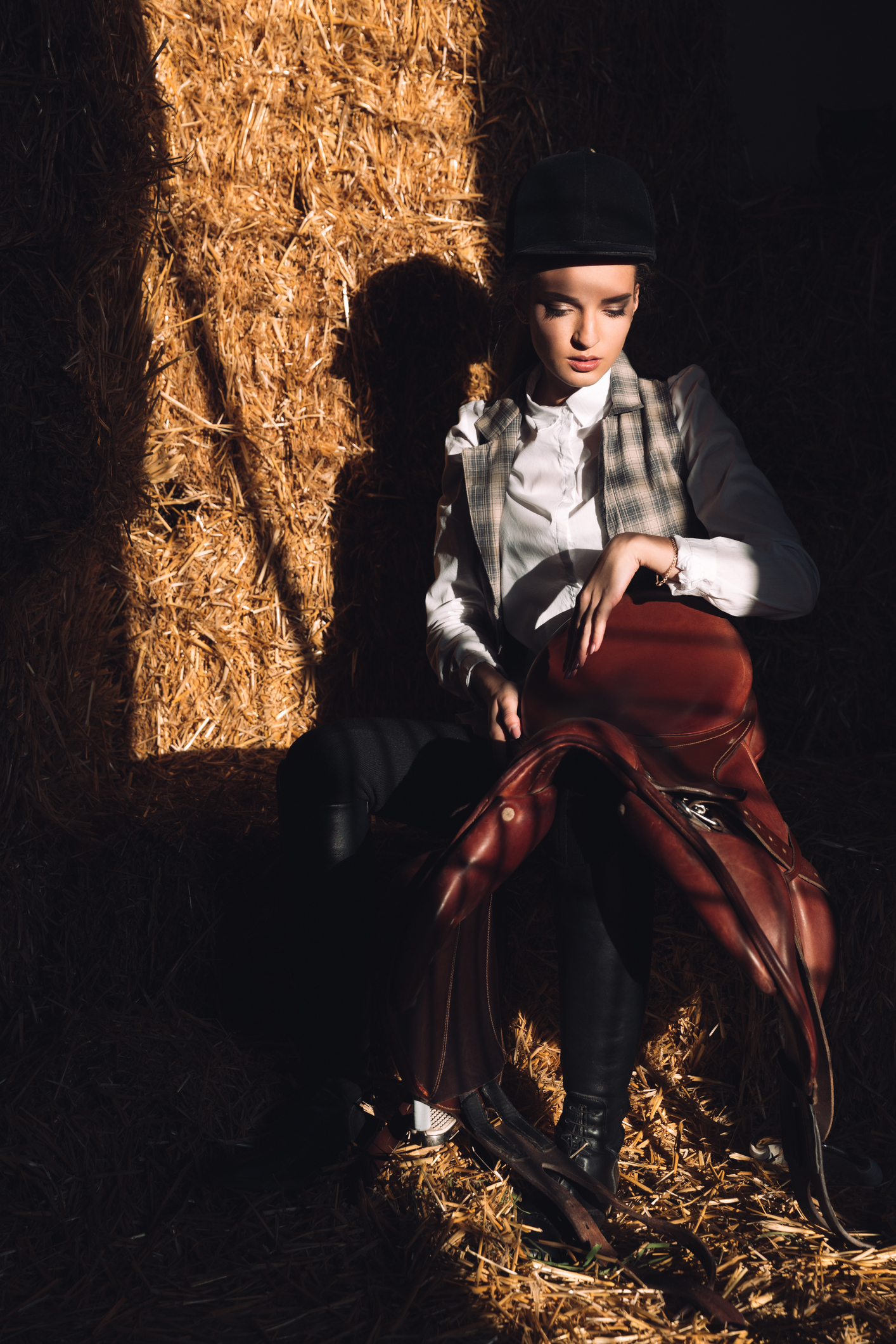 Image of serious young girl sitting in barn with seddle. Looking aside